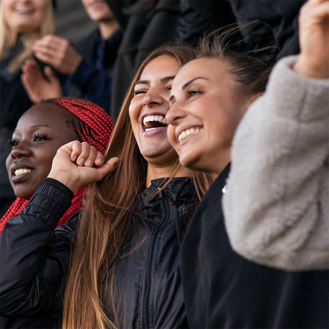Tres mujeres sonrientes celebran en un ambiente animado. Una de ellas tiene el cabello largo y castaño, otra lleva trenzas rojas, y la tercera usa una chaqueta clara. Todas parecen disfrutar de un momento de alegría y camaradería.