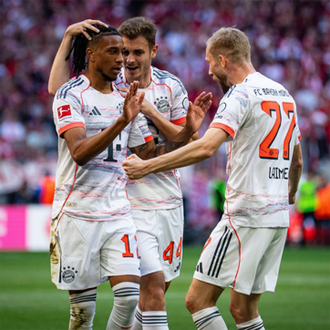 Celebración de tres jugadores del Bayern Múnich en un partido de fútbol. Están sonrientes y parecen emocionados después de un gol. Uno de ellos se lleva la mano a la cabeza, mientras otro aplaude. Visten camisetas blancas con detalles en naranja y negro.