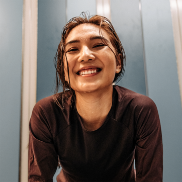 Una mujer sonriente con el cabello mojado lleva una camiseta de manga larga oscura. Su expresión refleja alegría y satisfacción, mientras se encuentra en un ambiente interior con paredes de color azul claro.