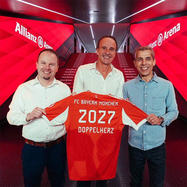 Tres hombres sostienen una camiseta del FC Bayern Múnich en un pasillo de la Allianz Arena. La camiseta es de color rojo con el número 2027 y la palabra "DOPPELHERZ". El ambiente es moderno, con luces y decoraciones en tonos rojos.