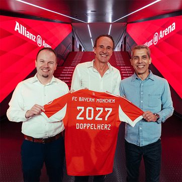 Tres hombres sostienen una camiseta del FC Bayern Múnich en un pasillo de la Allianz Arena. La camiseta es de color rojo con el número 2027 y la palabra "DOPPELHERZ". El ambiente es moderno, con luces y decoraciones en tonos rojos.